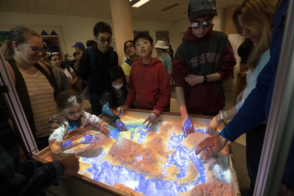 People standing around a sandbox with a colorful projector overlay.