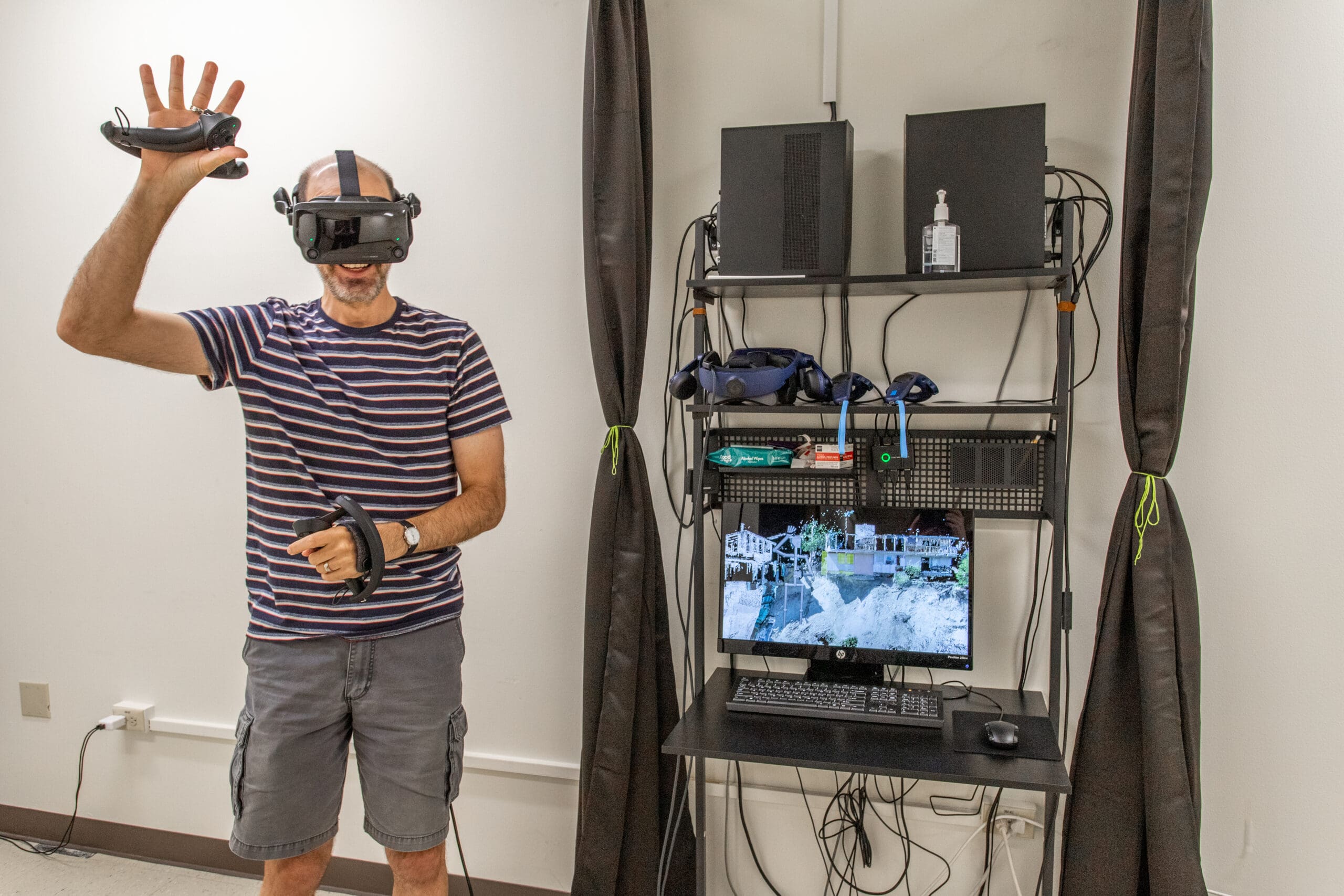 Image of a room with white walls, black tied back curtains, and a black rack with computer equipment. A data scientist in a striped shirt, khaki shorts, and VR headset stands with his arm upraised holding a controller.